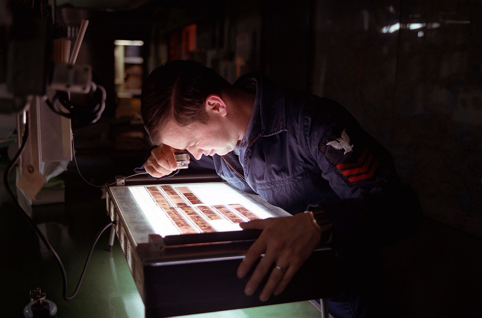 A photographer leans over a light table and checks a strip of negatives through a loupe